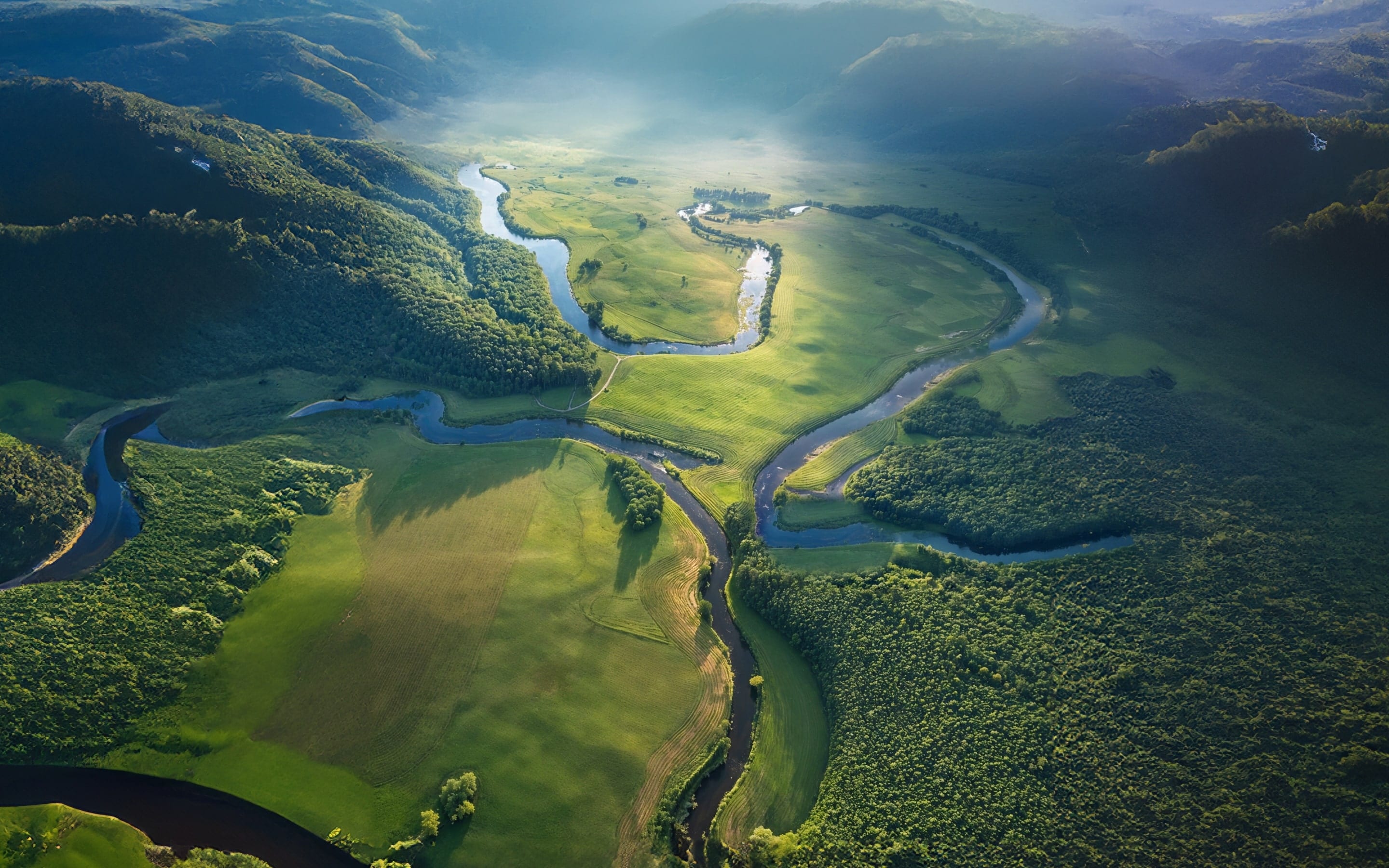 Aerial view of a meandering river flowing through lush green landscapes, surrounded by hills and forests, with soft light creating a serene atmosphere.